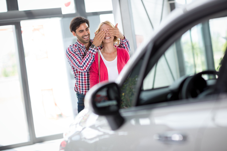 Couple stand in front of new car, man holding his wife covered her eyes